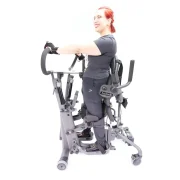 Eine Frau steht im EasyStand Glider. Seitenansicht: Linker Arm vorne, linkes Bein hinten; rechter Arm hinten, rechtes Bein vorne.