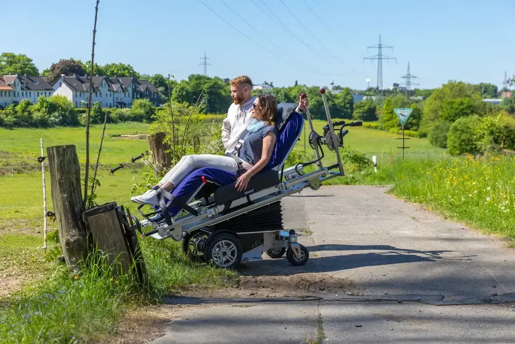 Ein Begleiter steht mit einer Frau, die im COSEATadapt fly liegt am Rand eines Feldwegs. Durch die angewinkelte Liegeposition und den nach vorne geneigten COSEAT ist sie fast auf der gleichen Augenhöhe, wie der Begleiter.