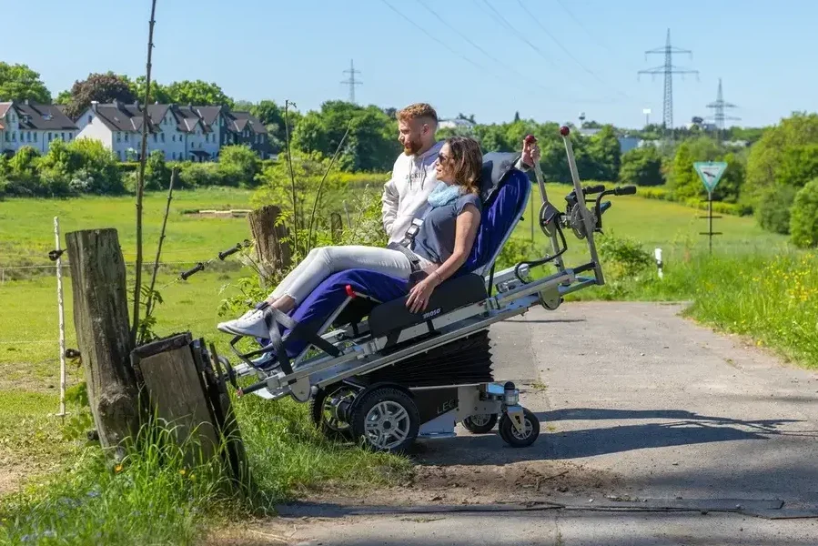 Ein Begleiter steht mit einer Frau, die im COSEATadapt fly liegt am Rand eines Feldwegs. Durch die angewinkelte Liegeposition und den nach vorne geneigten COSEAT ist sie fast auf der gleichen Augenhöhe, wie der Begleiter.